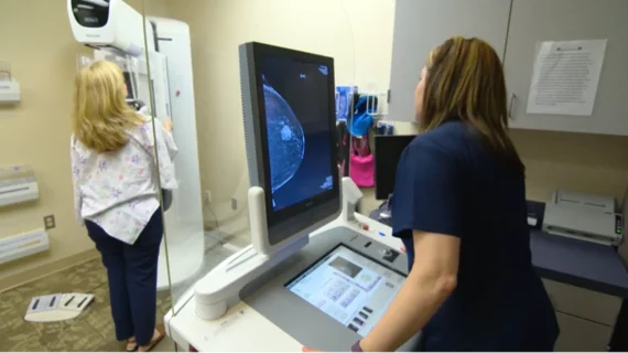 A mammography exam in progress with the patient on the breast imaging system and the technologist acquiring images. The image on the screen shows a suspect lesion.